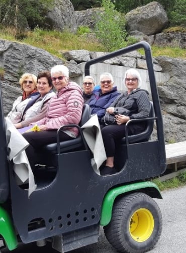 Six women smiling while riding in an open-air, green utility vehicle on a paved path near a rocky hillside.