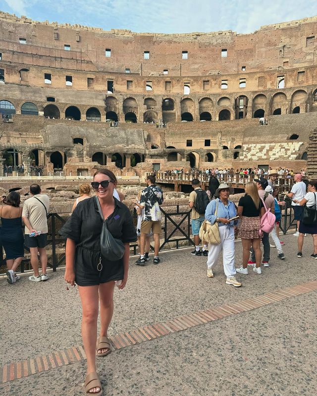 A woman in a black romper and sunglasses smiles while walking through the interior of the Roman Colosseum