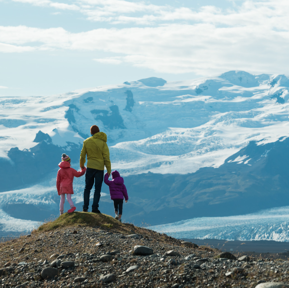 A man in a yellow jacket holds the hands of two children as they stand on a rocky hill