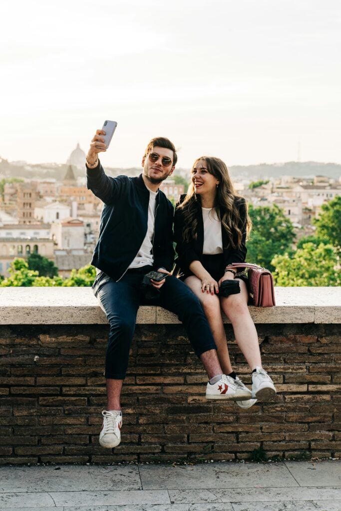 A young man and woman sitting on a stone ledge, smiling as they take a selfie together with a smartphone against a blurred European cityscape at sunset.