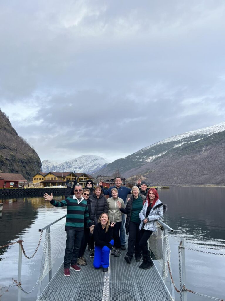 A group of ten travellers in winter coats poses on a metal pier extending over a calm lake