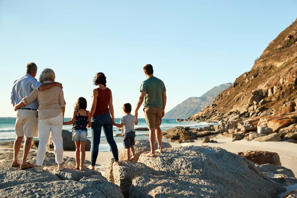 A multi-generational family of six stands on large beach rocks, looking out at the ocean and a rugged mountain coastline under a clear blue sky.