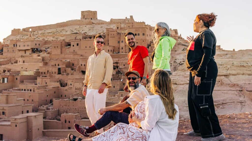 A diverse group of six friends smiles and talks while posing on a rocky overlook, with the ancient earthen buildings of Aït Benhaddou, Morocco, rising in the background.