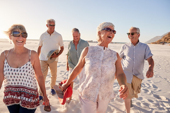 A group of five smiling senior friends walks together along a sunny, white sand beach under a clear blue sky.